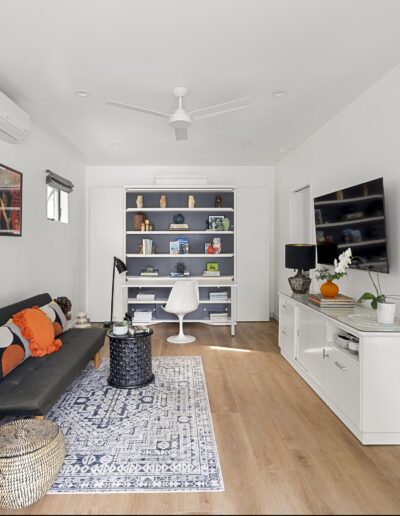 A modern living room features a black sofa with orange and green pillows, a wall-mounted TV, a white shelving unit with books and decor, and a desk with a chair. A patterned rug and indoor plants add décor.