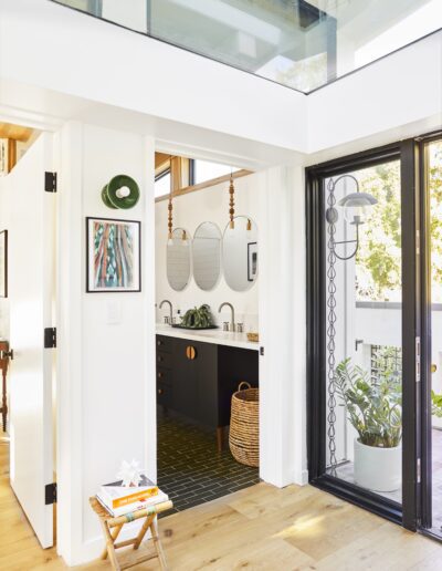 A modern bathroom with a black double vanity, two round mirrors, and a wicker basket. The space features natural light from large glass windows and a green plant near the doorway.